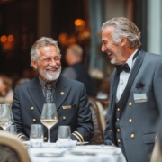 Two experienced hotel professionals in formal hospitality uniforms share a friendly conversation and laugh together beside a dining table in an elegant restaurant. The image highlights strong workplace relationships, leadership, mentorship, team culture, and employee engagement within the hospitality industry.